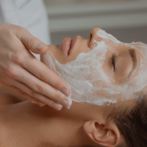 Cursos limpeza de pele Mulher deitada com olhos fechados. Sobre toda sua face há um creme branco e uma mão que massageia seu rosto.