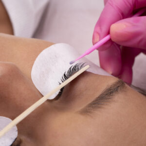 Aplicação de Cílios Profissional aplica extensão de cílios em cliente de olhos fechados, usando luvas rosas e pincel delicado, em ambiente de estética identificado como “Escola dos Cílios”.