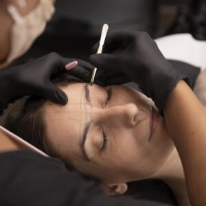 Design de Sobrancelhas Mulher deitada com olhos fechados. Acima de sua face aparecem duas mãos com luvas pretas, sendo que uma delas está segurando uma pinça profissional.